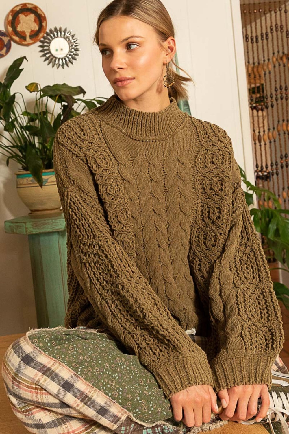 A person sits on a patterned blanket, wearing the POL Mock Neck Cable Knit Sweater and patterned pants, with plants and decorative items in the background.