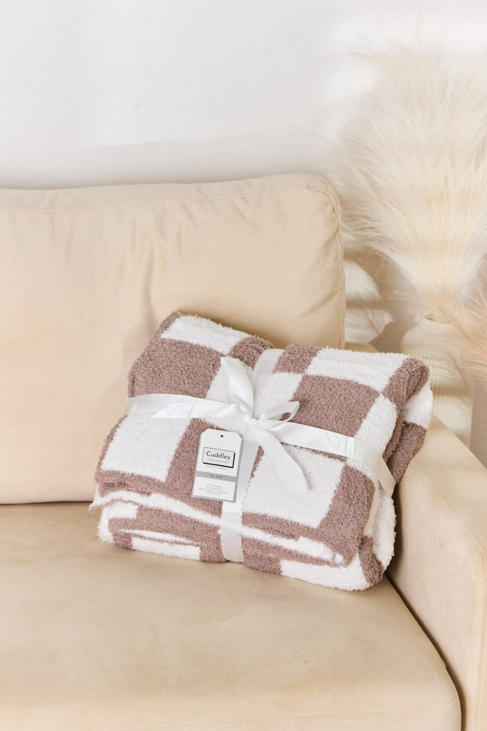 A person stands holding a large Cuddley Checkered Decorative Throw Blanket, smiling, in a room with a sofa and a potted plant. The blanket features a black and white checkerboard pattern, is made from 100% polyester, and imported for quality.