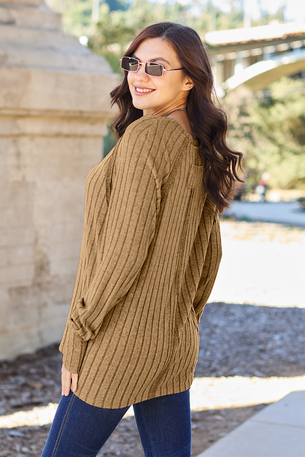 A person with long dark hair and sunglasses stands outdoors, wearing the Basic Bae Full Size Ribbed Round Neck Long Sleeve Knit Top in blue and a pair of jeans. The basic style outfit blends seamlessly with the background of trees and a bridge.