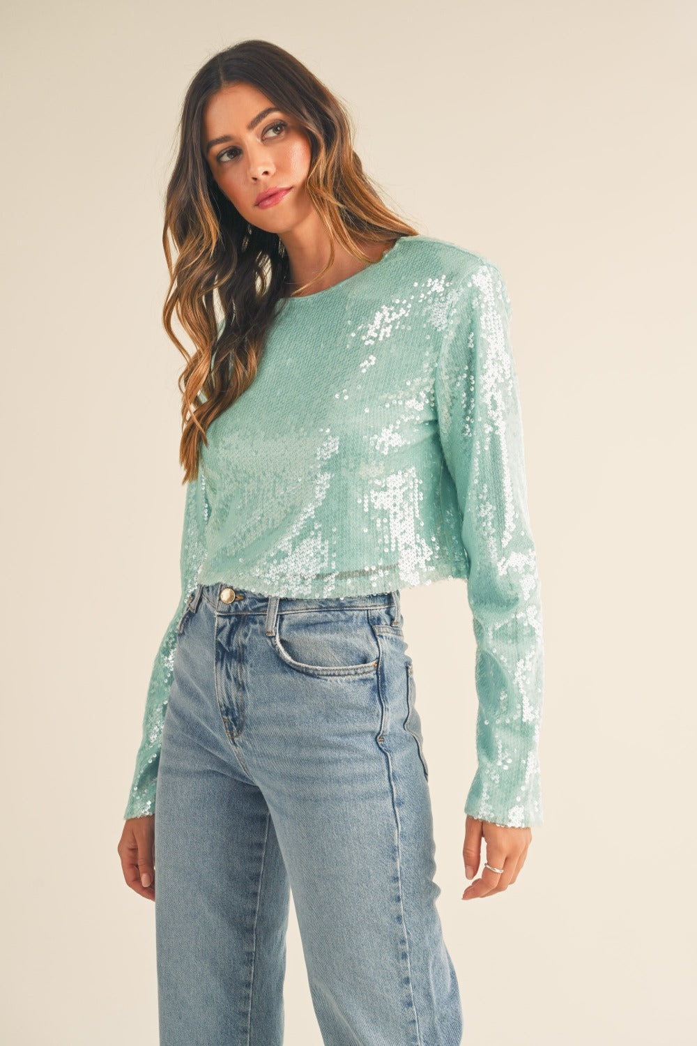 A person with long hair, wearing a fashion-forward, seafoam green MABLE Shoulder Padded Sequin Crop Top and high-waisted blue jeans, stands against a plain background.