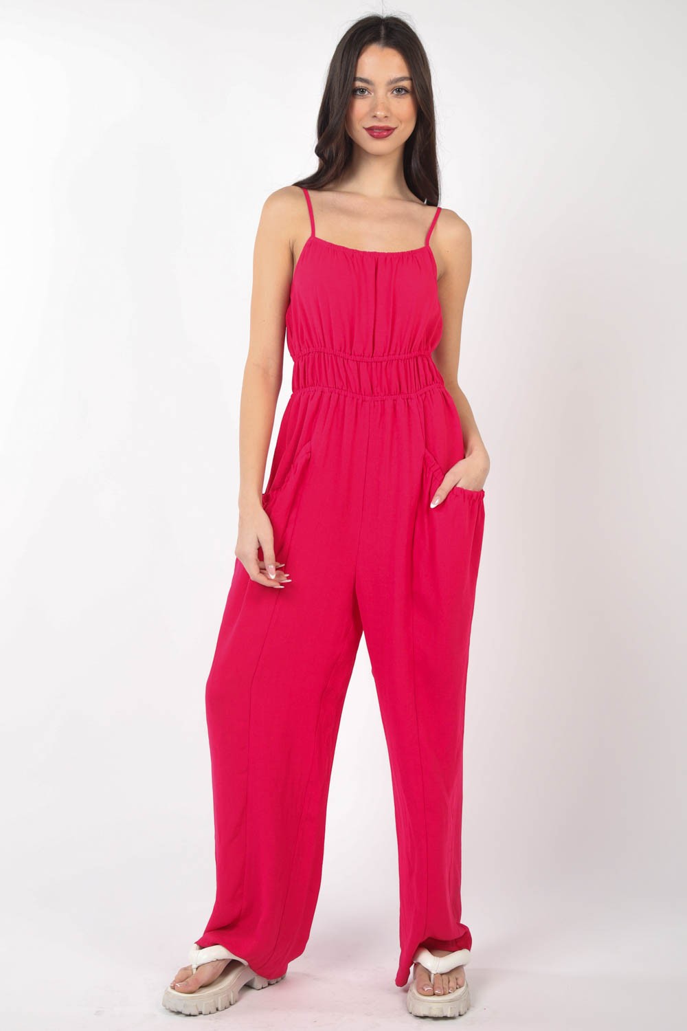 A person wearing the VERY J Pintuck Detail Woven Sleeveless Jumpsuit in bright pink paired with white sandals stands against a white background, with one hand placed in a pocket.