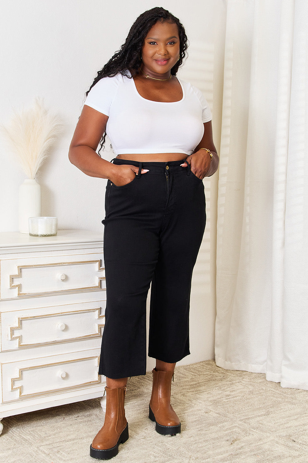 A person wearing Judy Blue Full Size High Waist Wide Raw Hem Cropped Jeans in black and a white lace top stands next to a white dresser adorned with a vase and candle.