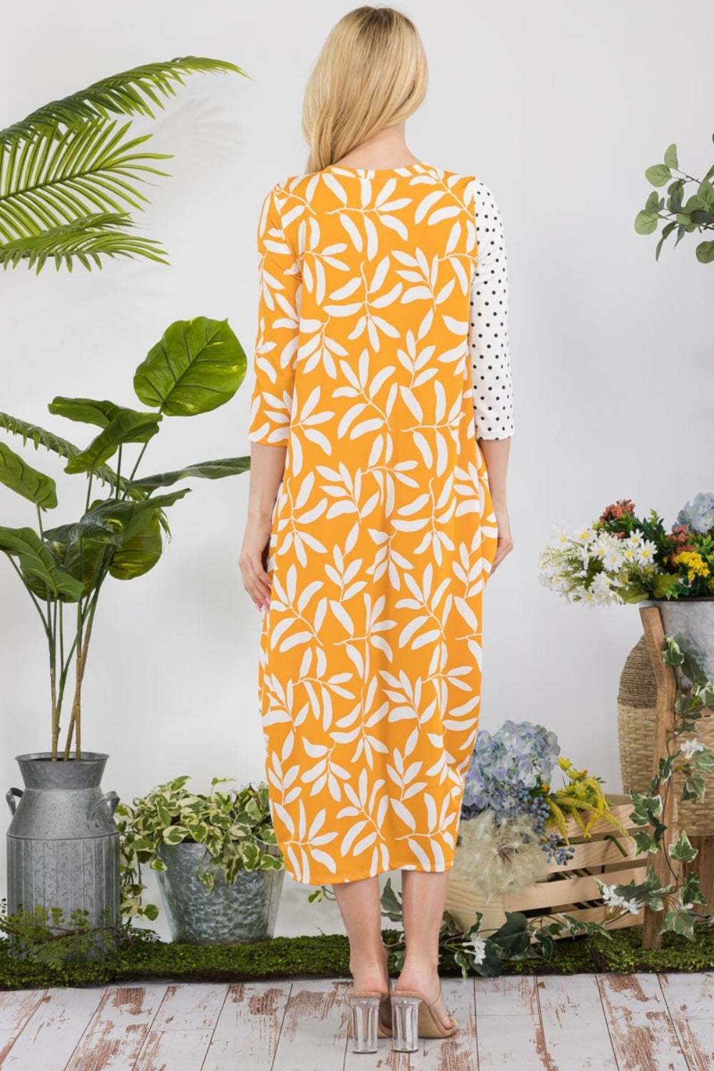 In a garden setting, a woman models the Celeste Full Size Floral Polka Dot Contrast Midi-Dress with Pockets, featuring its versatile yellow design among lush plants and vibrant blooms.