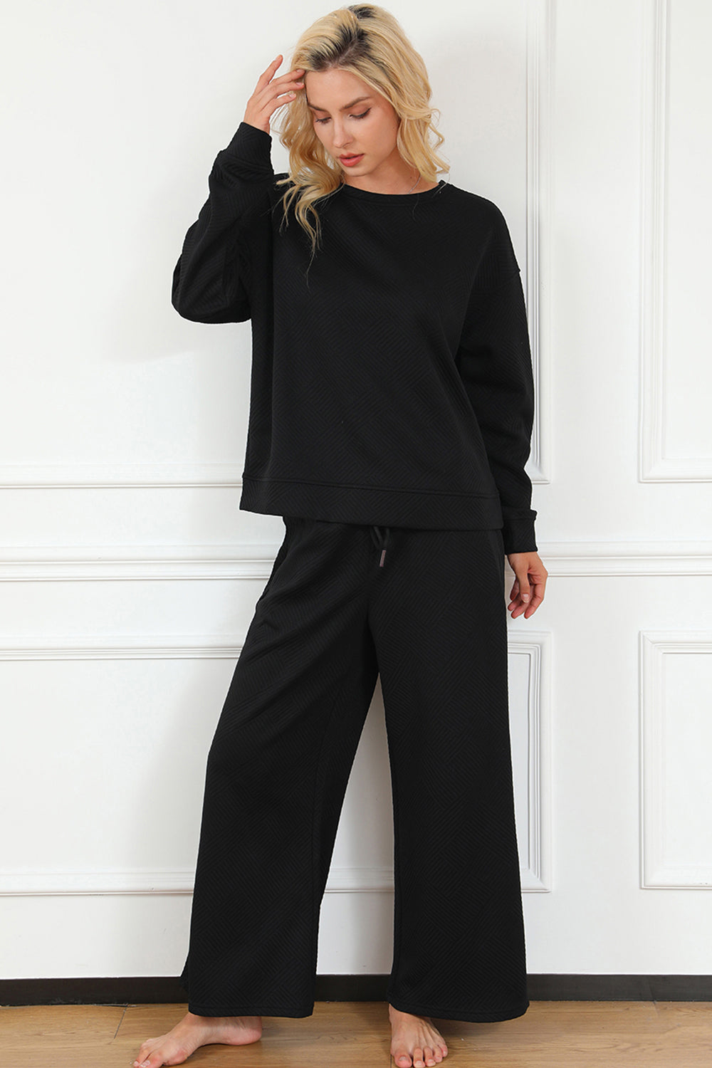 A woman with blonde hair, dressed in the Double Take Full Size Textured Long Sleeve Top and Drawstring Pants Set featuring a stretchy black long-sleeve top and matching wide-leg trousers made of rayon spandex, stands barefoot in a room with white walls and a wooden floor.