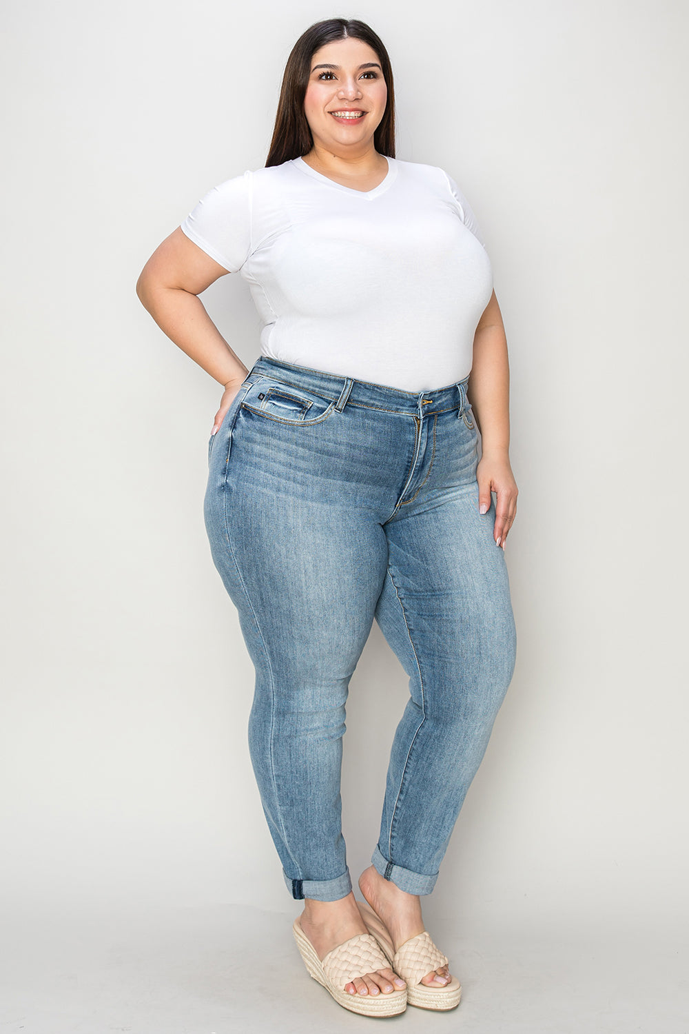 A person wearing a white shirt, Judy Blue Full Size Cuffed Hem Slim Jeans, and black wedge-heeled sandals is standing in a room with beige chairs and plants in the background.