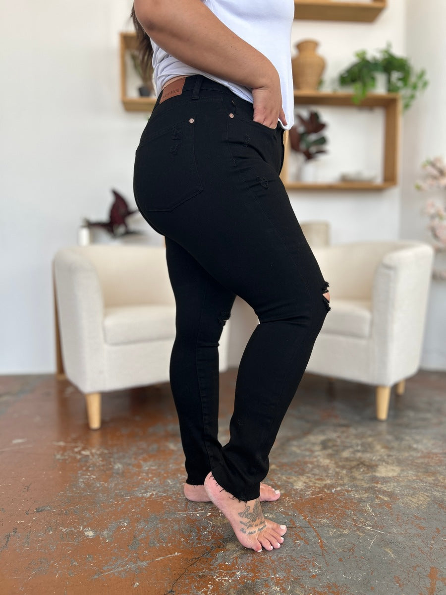 A person wearing a white T-shirt and Judy Blue Full Size Distressed Tummy Control High Waist Skinny Jeans poses barefoot in a room with white chairs and decorative plants in the background.