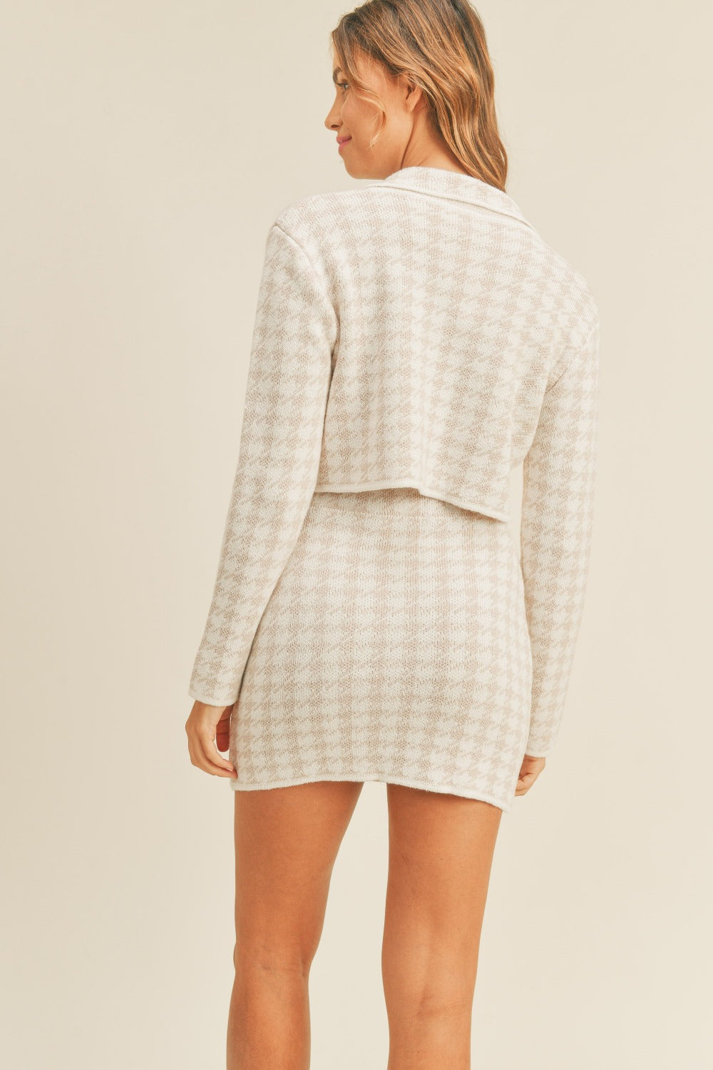 A person standing and wearing the MABLE Houndstooth Cropped Knit Cardigan and Mini Skirt Set, which features a chic aesthetic with a light-colored, long-sleeved button-up top paired with a matching mini skirt.