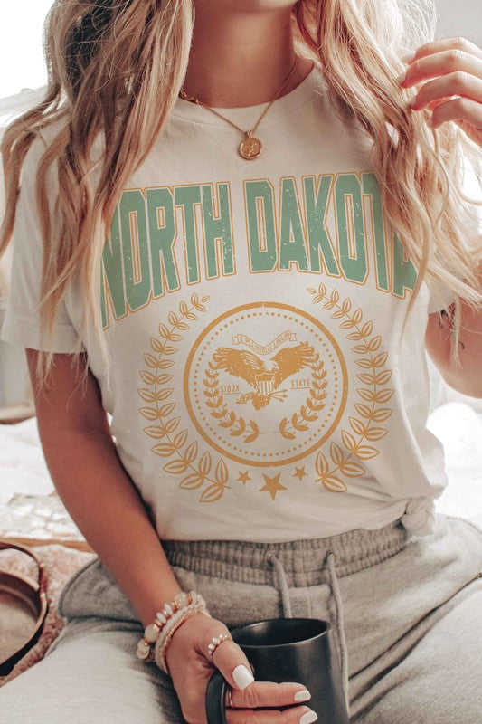 Person wearing a "NORTH DAKOTA Graphic Tee" featuring green and yellow text and graphics, holding a small object in their right hand.