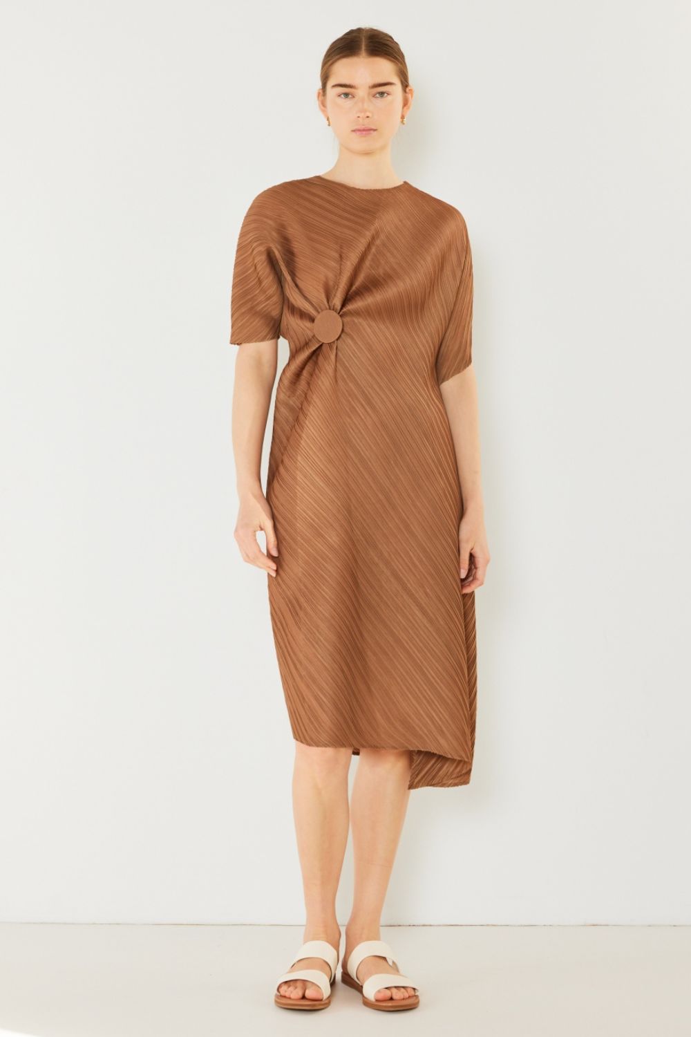 A woman stands against a plain backdrop wearing the stylish Marina West Swim Pleated Dolman Sleeve Dress, which features a knee-length, short-sleeved design in a textured brown fabric with an eye-catching circular detail at the waist. She completes her look with white sandals. This modern chic dress exudes versatility, making it perfect for various occasions.