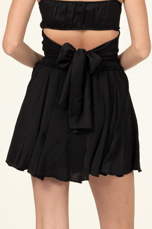 A person is partially visible in the image wearing the Not Your Girl Drawstring Ruffled Mini Skirt, featuring a bright pink flared silhouette with an elastic waistband and drawstring, paired with a matching top.