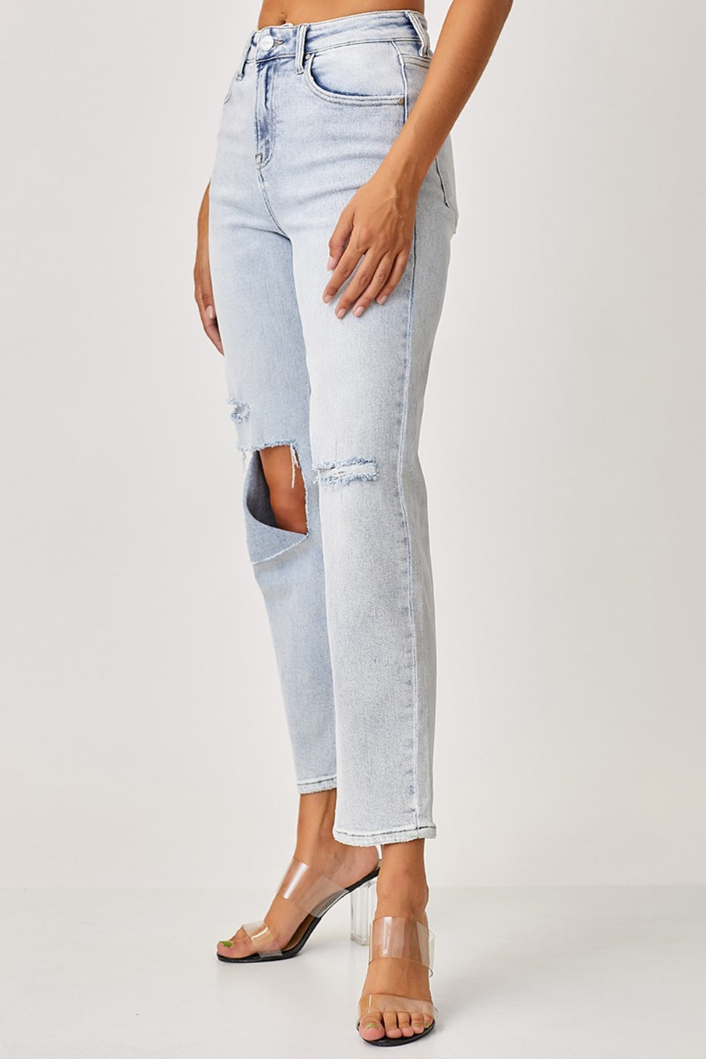 A person wearing RISEN High Rise Distressed Relaxed Jeans in light blue with a large knee tear and clear-heeled sandals.