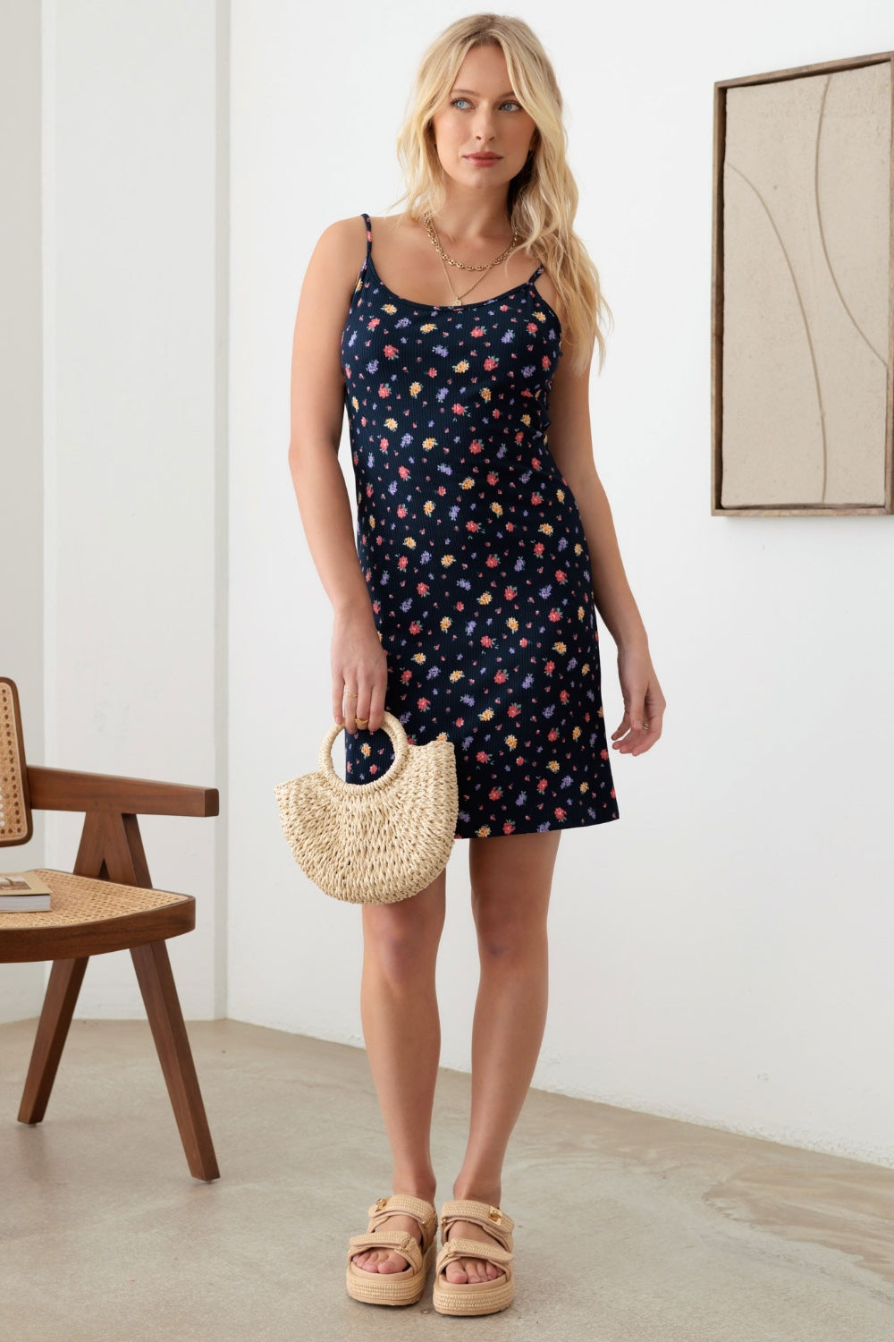 A woman with long blonde hair is wearing a Gilli Floral Ribbed Tie-Back Cami Mini Dress. She is holding a woven straw handbag and standing in a minimalist room.