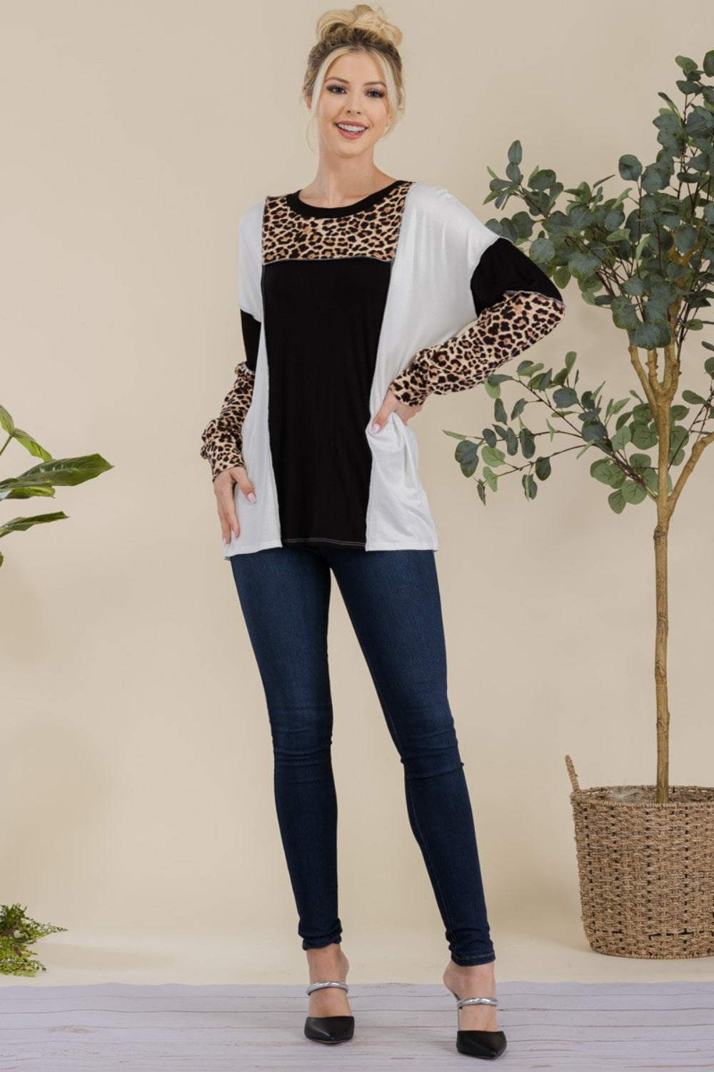 A woman stands in front of a simple background with a plant, wearing the Celeste Full Size Leopard Color Block Exposed Seam T-Shirt, which boasts leopard print cuffs and striking black and white color blocks.