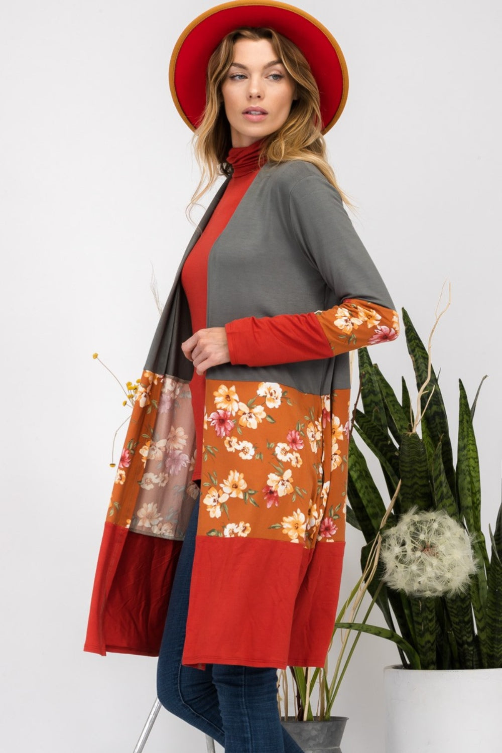 Wearing the Celeste Full Size Floral Color Block Open Front Cardigan, a person in a red hat stands indoors near potted plants, smiling.