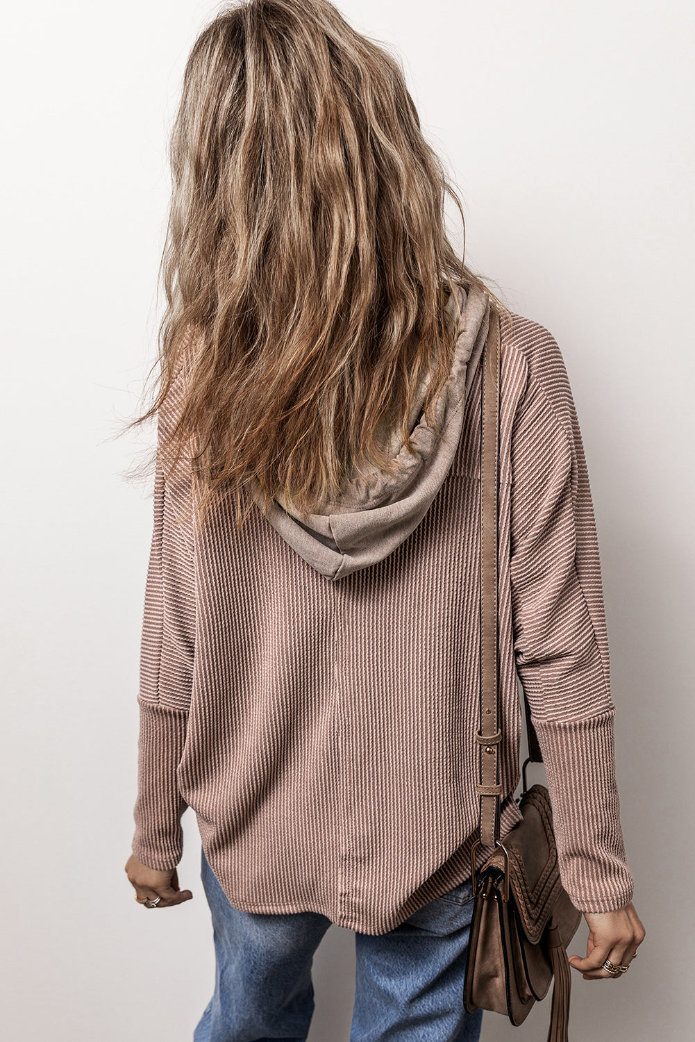 A person with long, wavy hair is facing away from the camera. They are wearing a Dark Brown Flap Pocket Patchwork Corded Drawstring Hoodie and blue jeans, holding a brown shoulder bag.