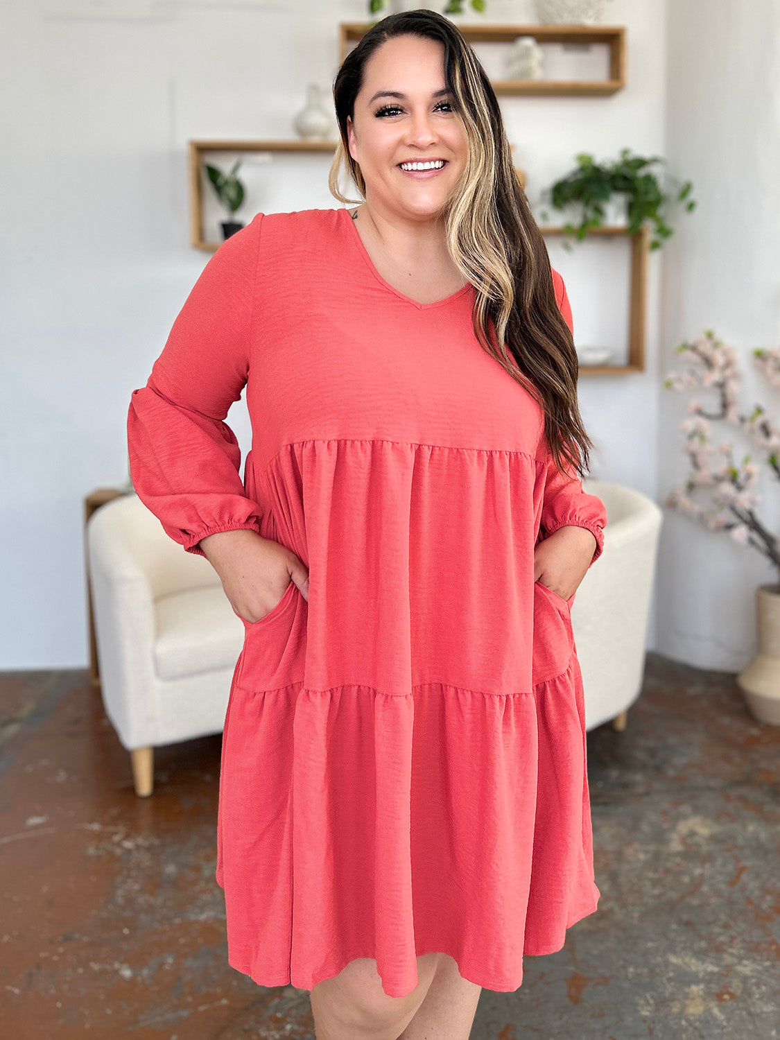 A woman stands wearing the Double Take Full Size V-Neck Balloon Sleeve Tiered Dress with Pockets in pink. Crafted from high stretch yarn, this dress combines comfort and style effortlessly. She has long, wavy hair and accessorizes with hoop earrings and a necklace. This imported garment is easy to care for – simply machine wash cold to keep it looking fresh.