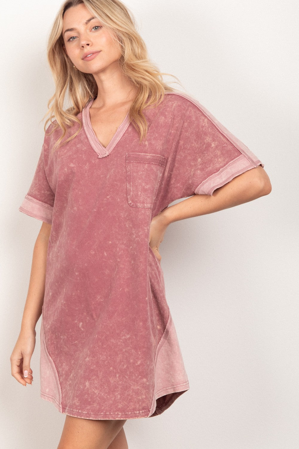 A woman stands against a plain background, wearing the loose-fitting, pink VERY J Short Sleeve V-Neck Tee Dress. Exuding casual style, she gazes at the camera with a slight smile while holding the side of her versatile piece.