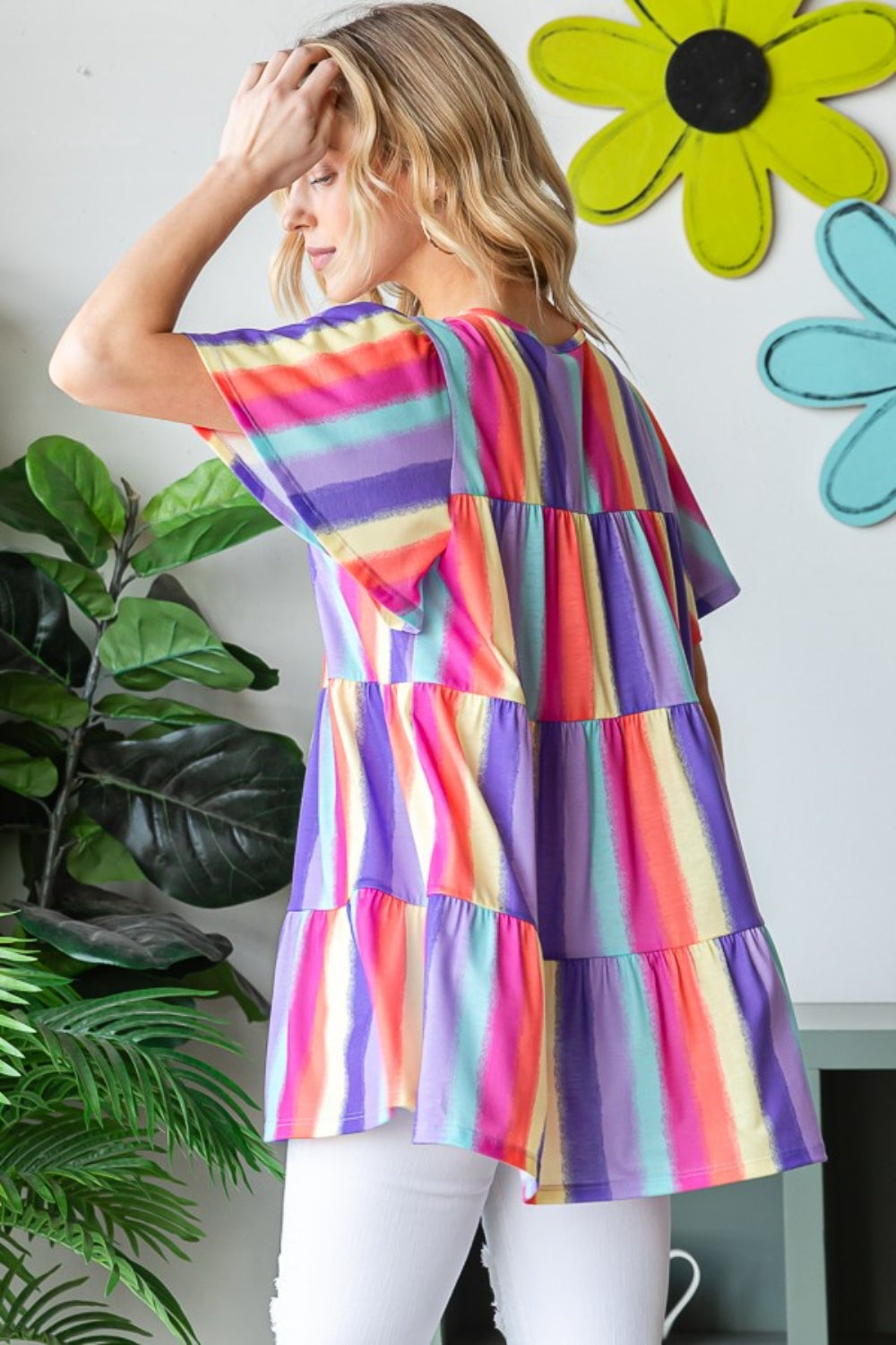 A person wearing the Heimish Full Size Short Sleeve Striped Tiered Top, featuring a colorful, classic striped pattern and flared sleeves, stands by a window. They are smiling, surrounded by plants in the background.