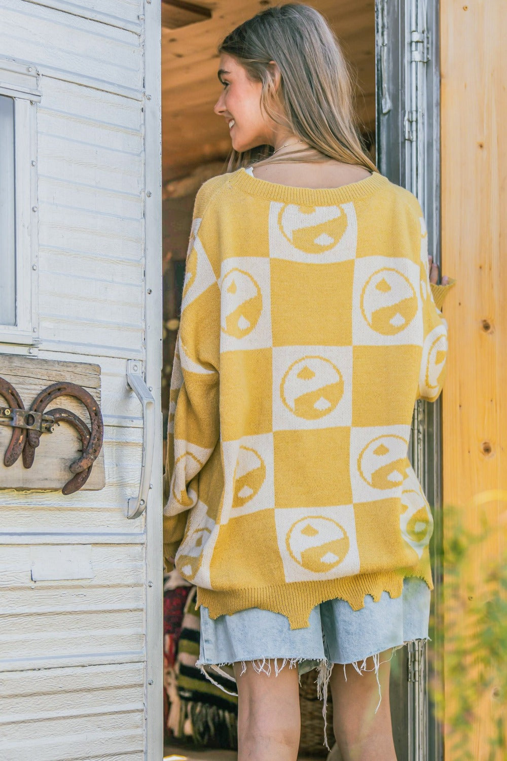 A woman wearing the And The Why Checkerboard Sweater with a Yin Yang pattern and distressed denim shorts stands in a doorway.