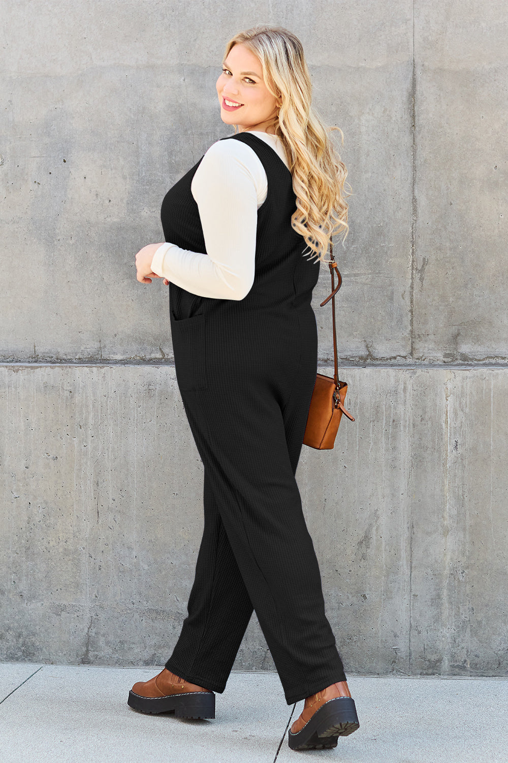 A person stands outdoors wearing the Double Take Full Size Sleeveless Straight Jumpsuit in blue and brown sandals, holding a cream-colored bag. In the background, there's a wooden bench and palm trees.