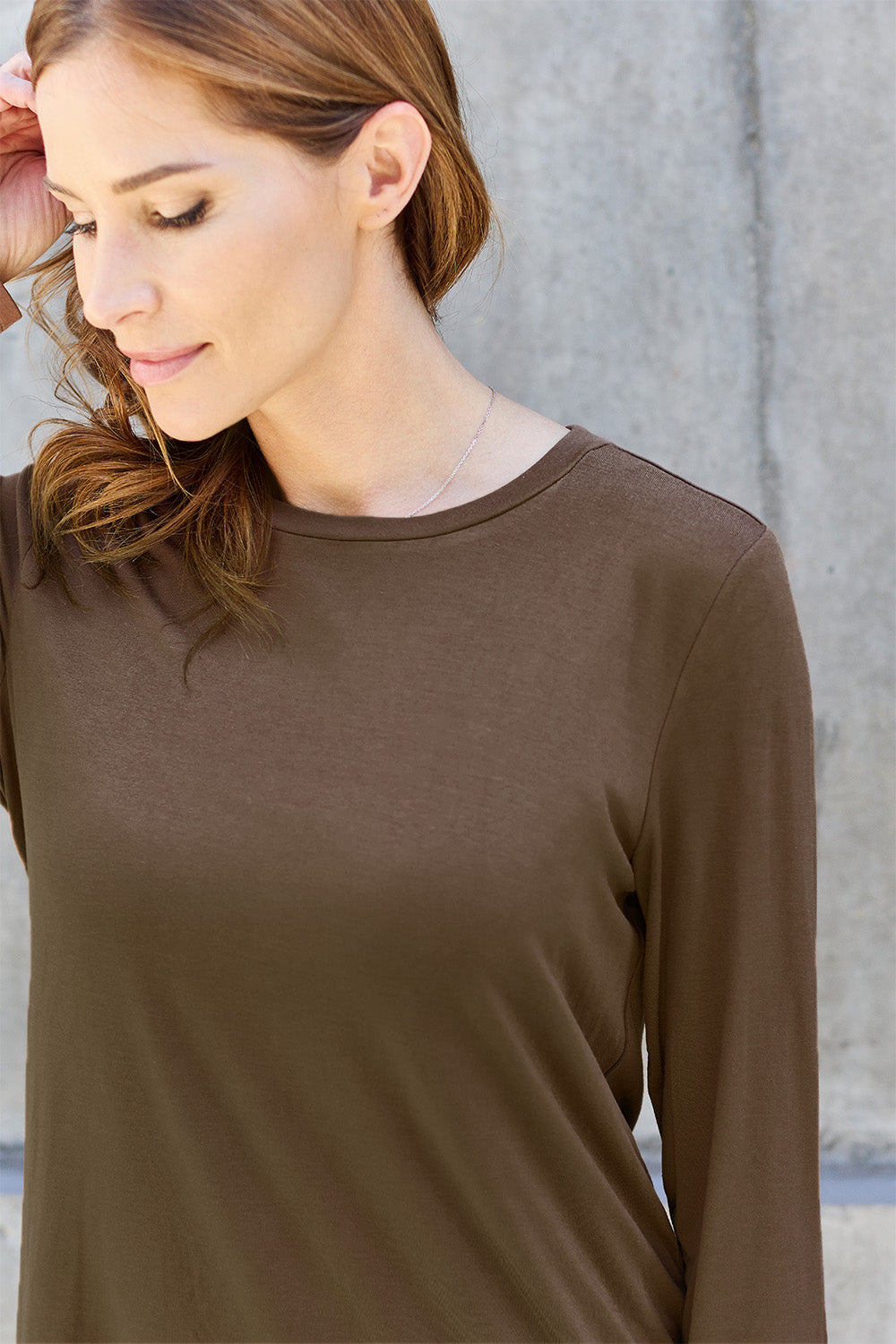 A woman with shoulder-length hair wearing the Basic Bae Full Size Round Neck Long Sleeve Top and blue jeans stands in front of a concrete wall, holding a beige handbag over her shoulder. Her outfit exemplifies basic style, with the top made from 100% polyester and recommended for machine wash cold.