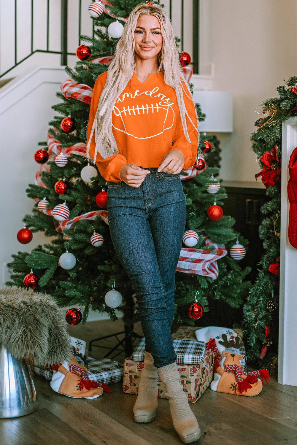 A woman with blonde hair, wearing an Orange Game Day Lettering Rugby Football Notched Neck Sweatshirt and blue jeans, stands next to a decorated Christmas tree.