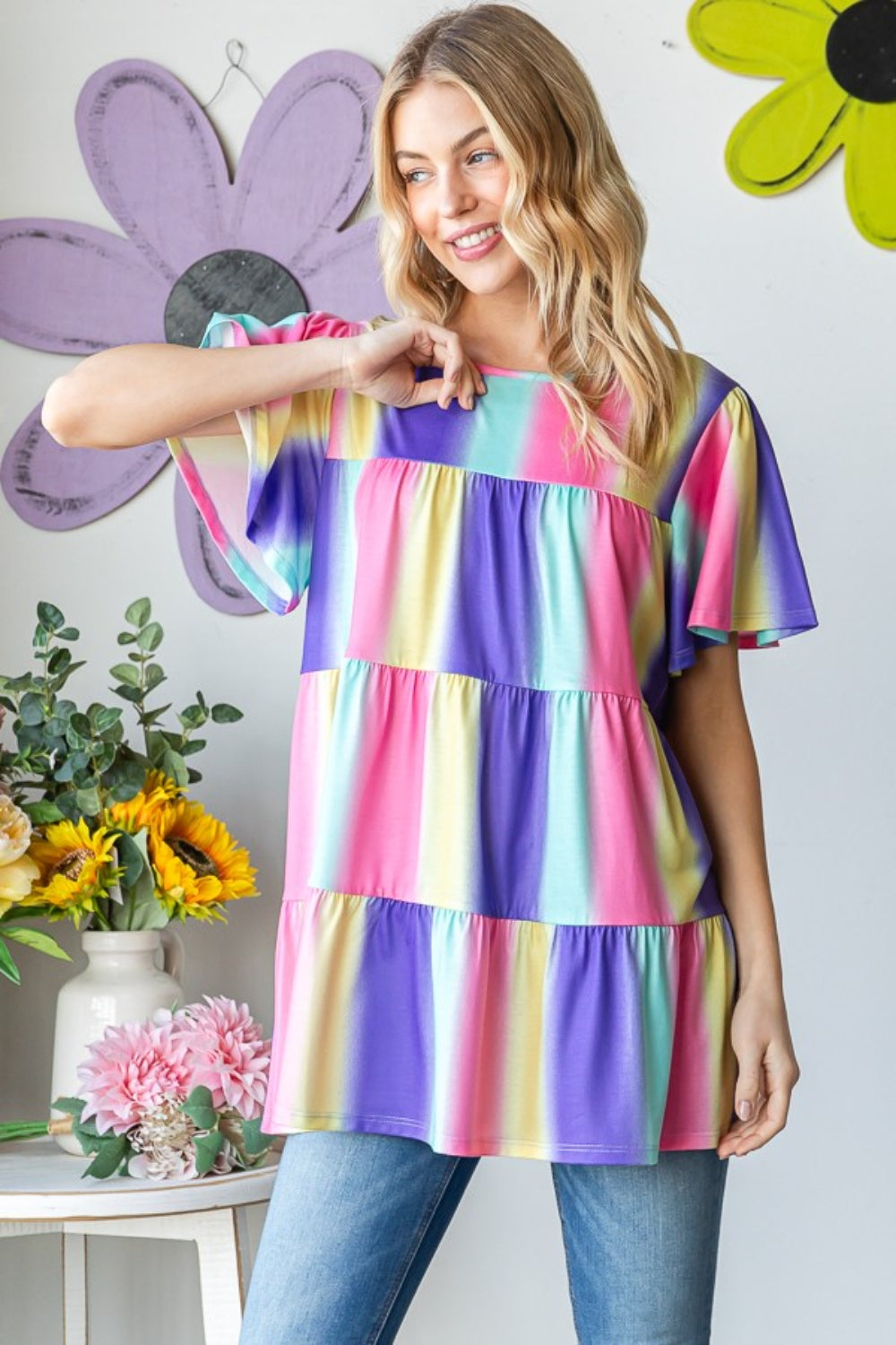 A person poses in a striking Heimish Full Size Short Sleeve Striped Tiered Top, surrounded by decorative flowers and wall accents.