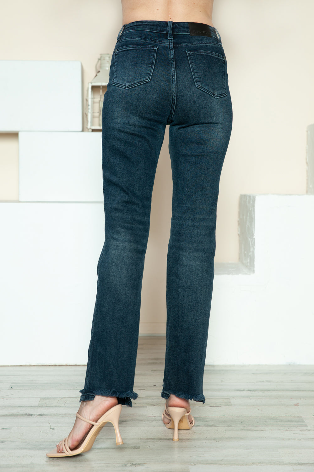 A person is modeling the Judy Blue Full Size Button Fly Hem Destroy Straight Jeans, high-waisted with distressed detailing and frayed hems, paired with light-colored open-toe heels. The background includes white blocks and a light-colored wall.