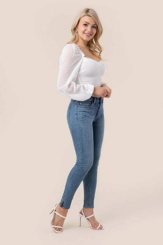 A person wearing the LS shirring sleeve bodysuit with Swiss dot mesh, featuring sheer, long puffed sleeves and a sweetheart neck, paired with light-colored jeans, stands against a plain background.