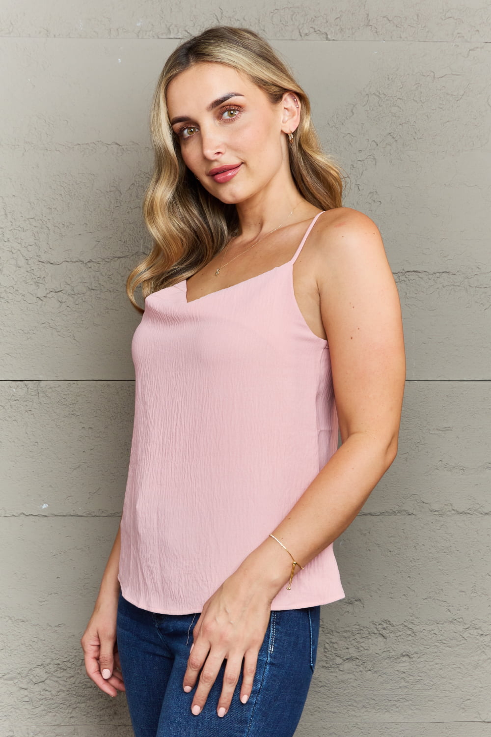 A person in a Ninexis For The Weekend Loose Fit Cami and jeans stands against a textured gray wall, smiling and looking to the side.