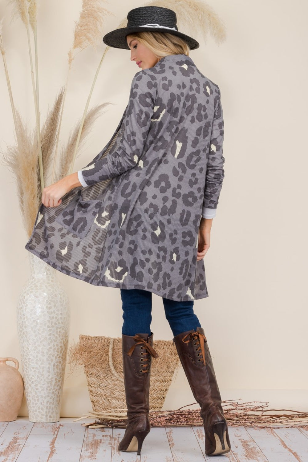 A person wearing a Celeste Full Size Leopard Open Front Contrast Cardigan over a white top and jeans, complemented by brown boots, accessorized with a black hat and necklace, stands next to a tall vase with decorative grasses.