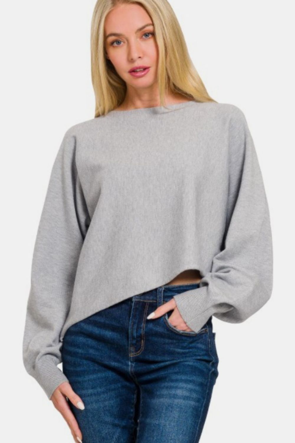 A person with long blonde hair wears a stylish Zenana Asymmetric Hem Long Sleeve Sweater made of soft and comfortable gray fabric, paired with blue jeans, standing against a plain white background.