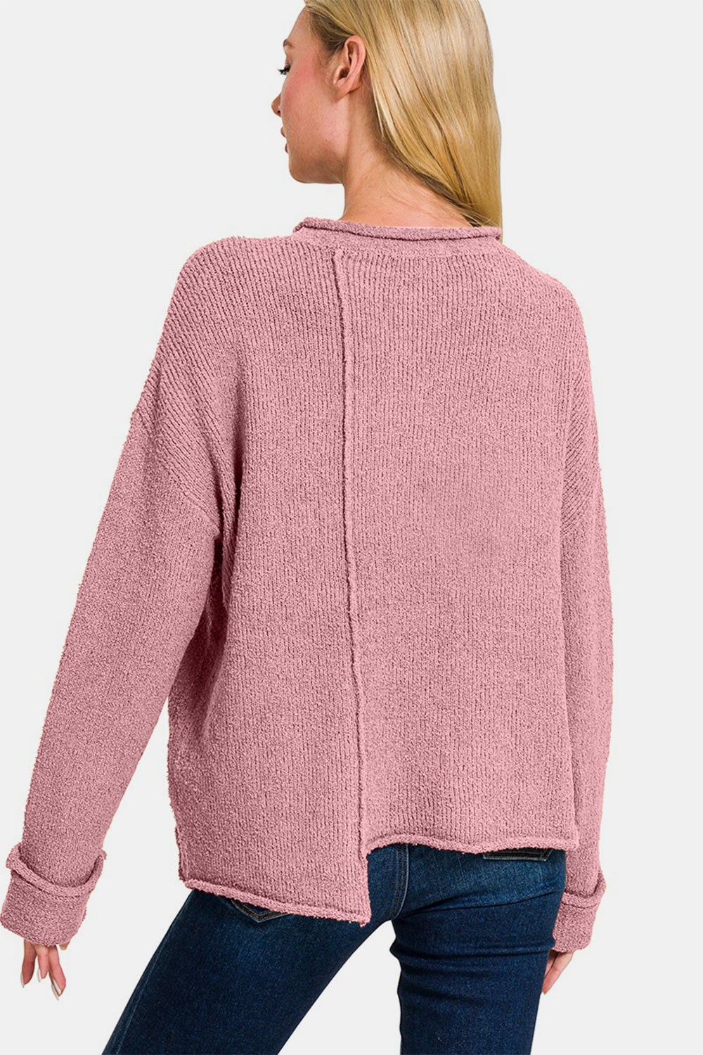 A person with long blonde hair is wearing a Zenana Asymmetric Hem Drop Shoulder Sweater in light pink and blue jeans, standing against a plain white background.