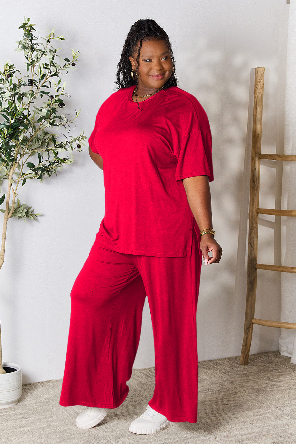 A woman stands near a potted plant and wooden ladder, wearing the Double Take Full Size Round Neck Slit Top and Pants Set in a bright pink two-piece outfit with matching wide-leg pants that are available in plus size and are easily machine washable.