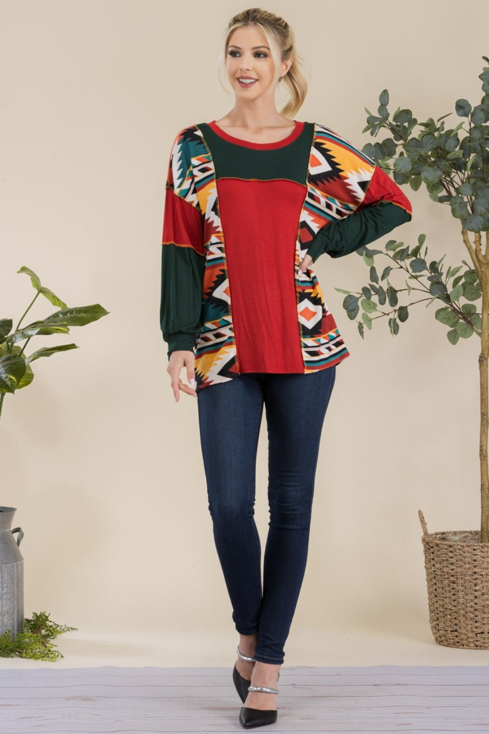 A woman stands against a plain background, wearing the Celeste Full Size Exposed Seam Printed Color Block T-Shirt with vibrant red, green, and yellow geometric sections, paired with dark jeans. She smiles, her hands resting in her jeans' pockets.