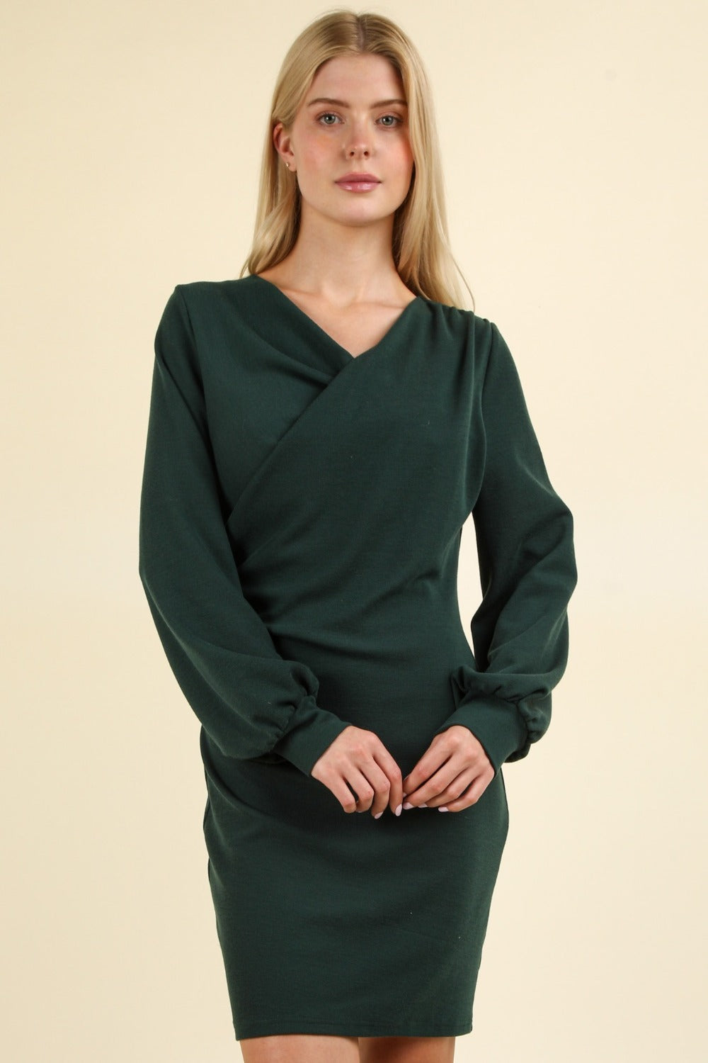 A person stands facing the camera, wearing the VERY J Ruched Detail Bodycon Mini Dress in dark green, featuring a V-neck and a wrap-style bodice. The figure-hugging silhouette accentuates their form beautifully, set against a plain, light-colored background.