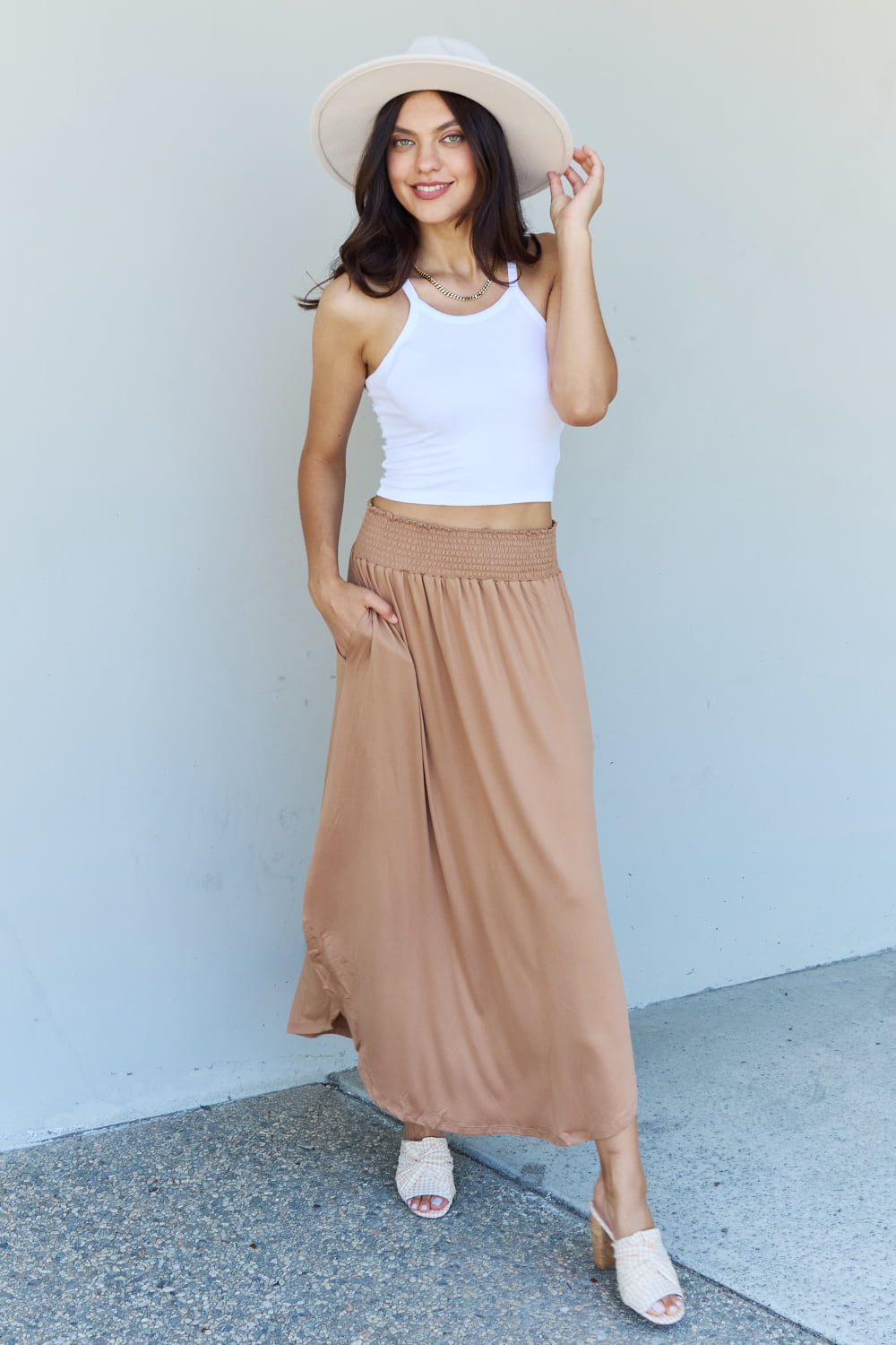 A person stands against a wall, wearing a white sleeveless top, the Doublju Comfort Princess Full Size High Waist Scoop Hem Maxi Skirt in Tan, and blue platform sandals. The right hand rests on the side of the skirt.