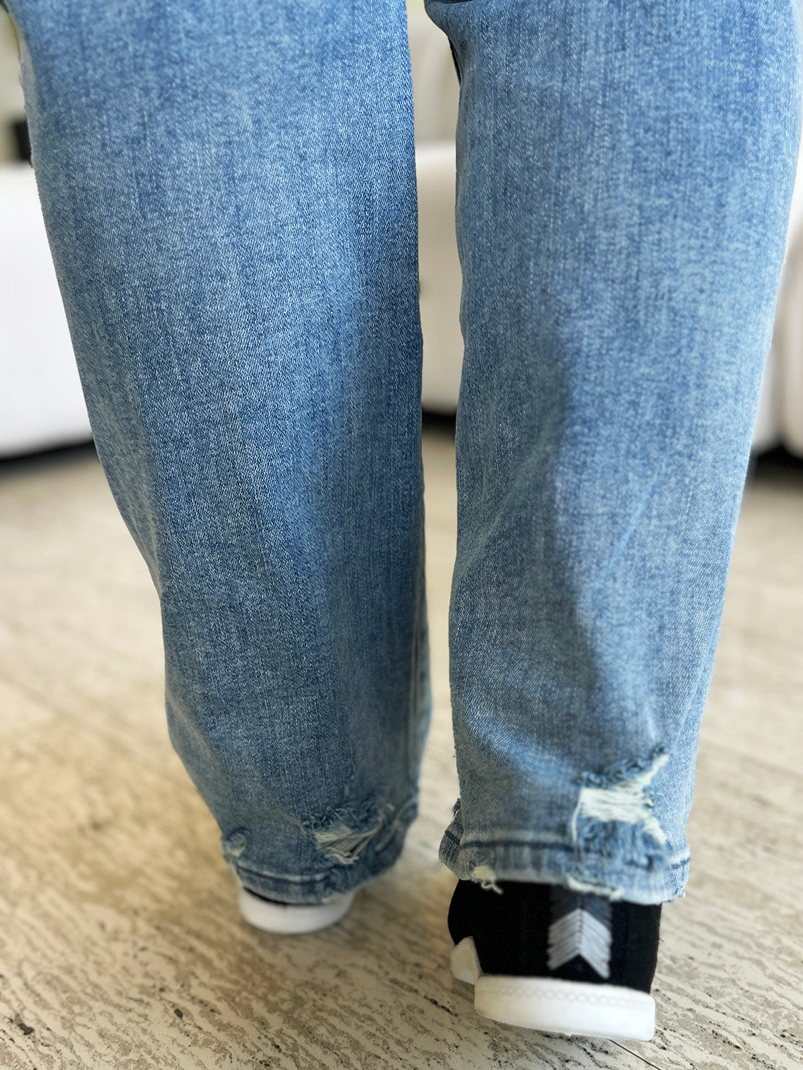 A person wearing Judy Blue Full Size Mid Rise Distressed Straight Jeans in light blue with a white top and white slip-on shoes stands in a room with a white couch and a plant.