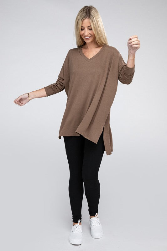 A woman with long, blonde hair is wearing a loose-fitting, brown Dolman Long Sleeve V-Neck Side Slit Hi-Low Hem Top and black pants. She poses with one hand on her hip and a neutral facial expression against a light grey background.