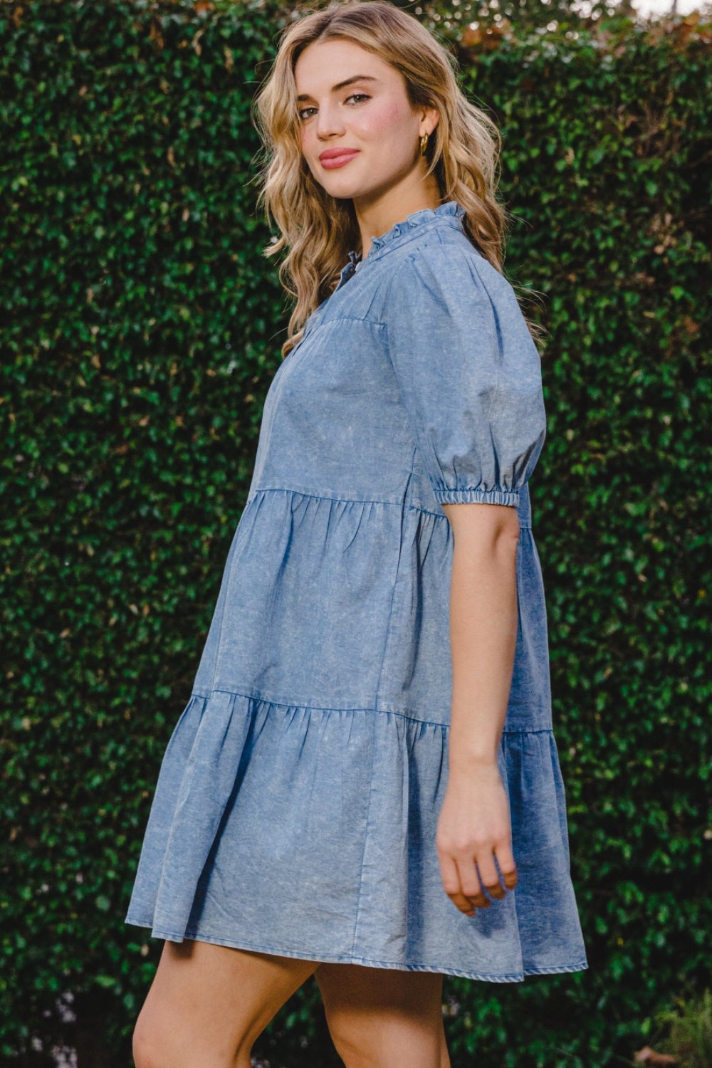 A person wearing the ODDI Washed Tiered Mini Denim Dress with puffed sleeves stands in front of a green leafy background.