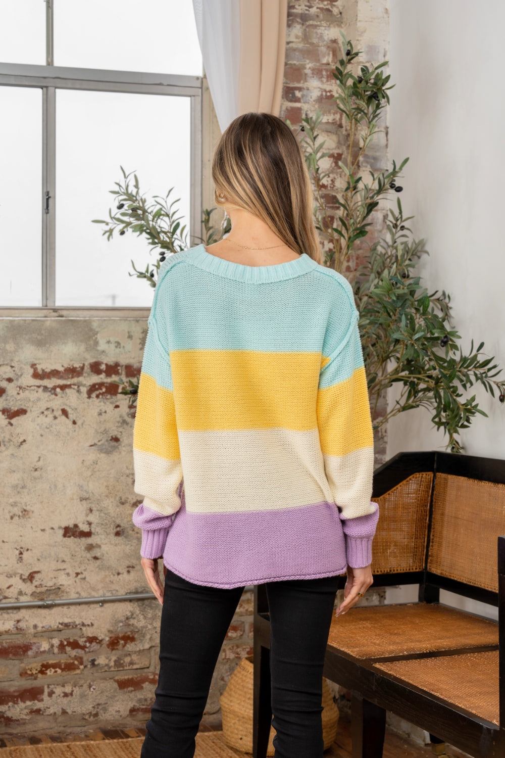A person stands indoors near a window, wearing the Sew In Love Full Size Color Block Exposed Seam Sweater and black pants. The cozy knitwear complements the rustic walls and nearby plant, adding a touch of warmth to the scene.