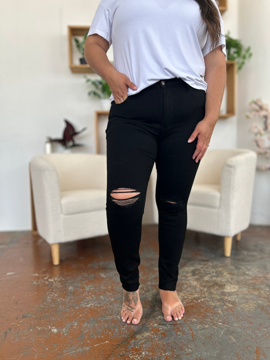 A person wearing a white T-shirt and Judy Blue Full Size Distressed Tummy Control High Waist Skinny Jeans poses barefoot in a room with white chairs and decorative plants in the background.