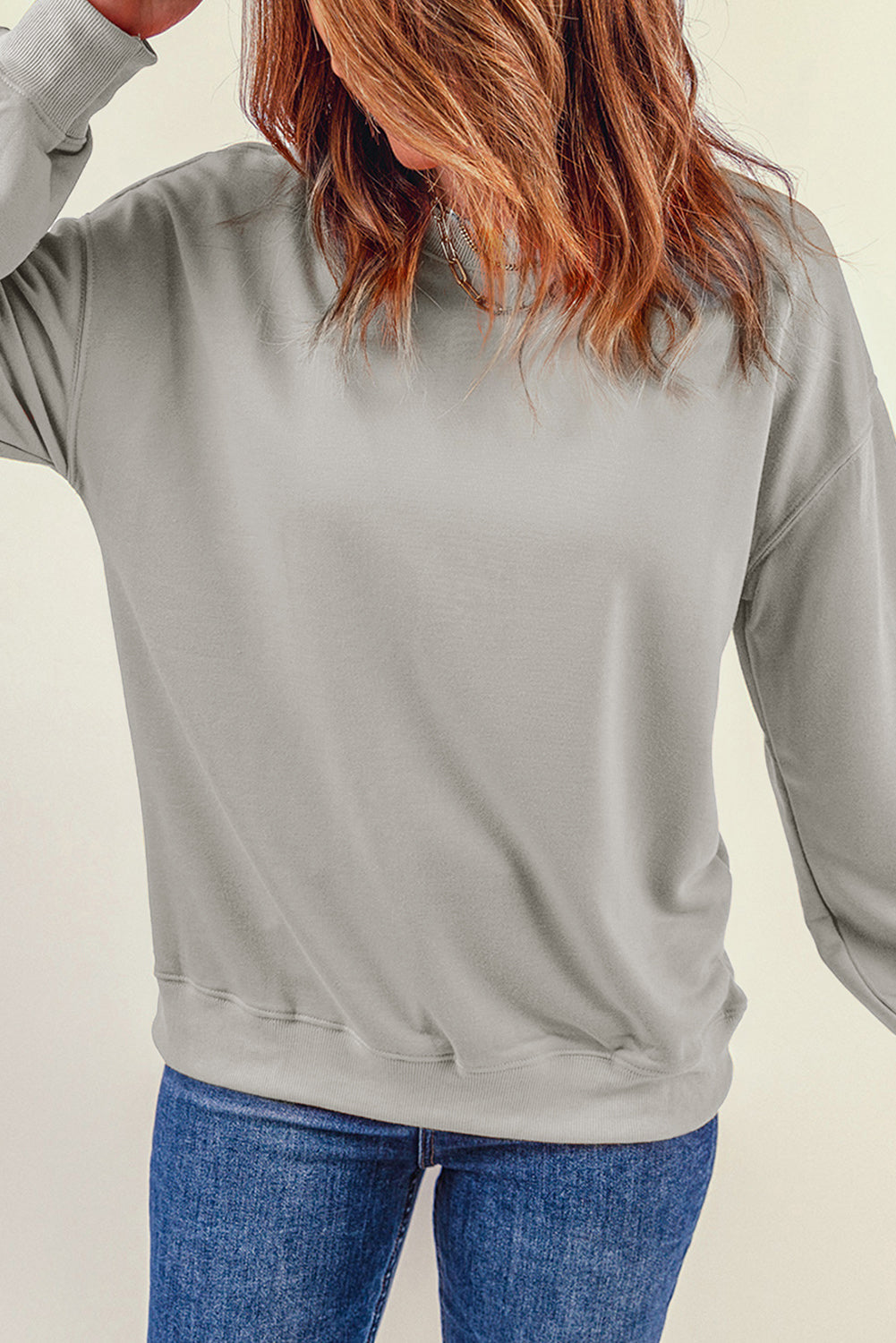 Person wearing a Gray Plain Crew Neck Pullover Sweatshirt and blue jeans with a hand in their pocket, adorned with layered necklaces.