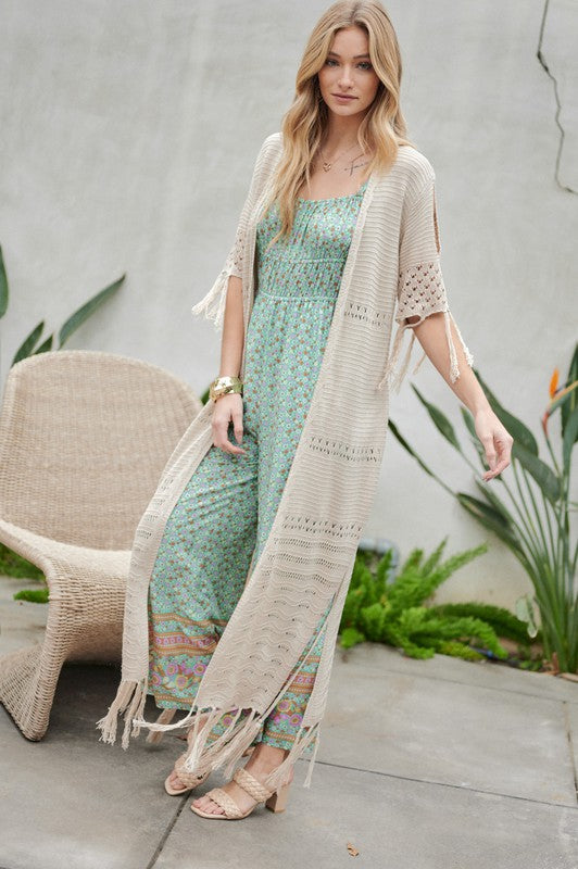 A woman in a green floral dress and a Solid Long Cardigan With Fringe exudes boho-chic vibes as she strolls on a patio, surrounded by outdoor furniture and lush plants.