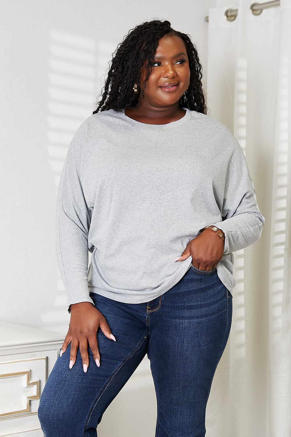 A person with long hair wearing a Double Take Seam Detail Round Neck Long Sleeve Top in solid black and blue jeans stands with one hand in their pocket, looking downward and smiling.