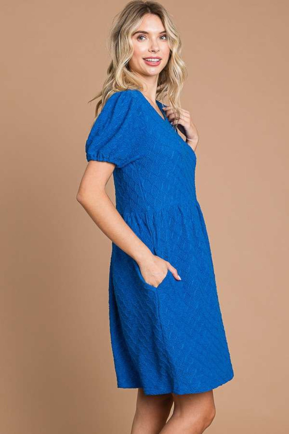 Against a plain brown backdrop, an individual is styled in a Culture Code Texture Round Neck Short Sleeve Dress with Pockets, exuding elegance and flair with its blue textured fabric and chic short puff sleeves.