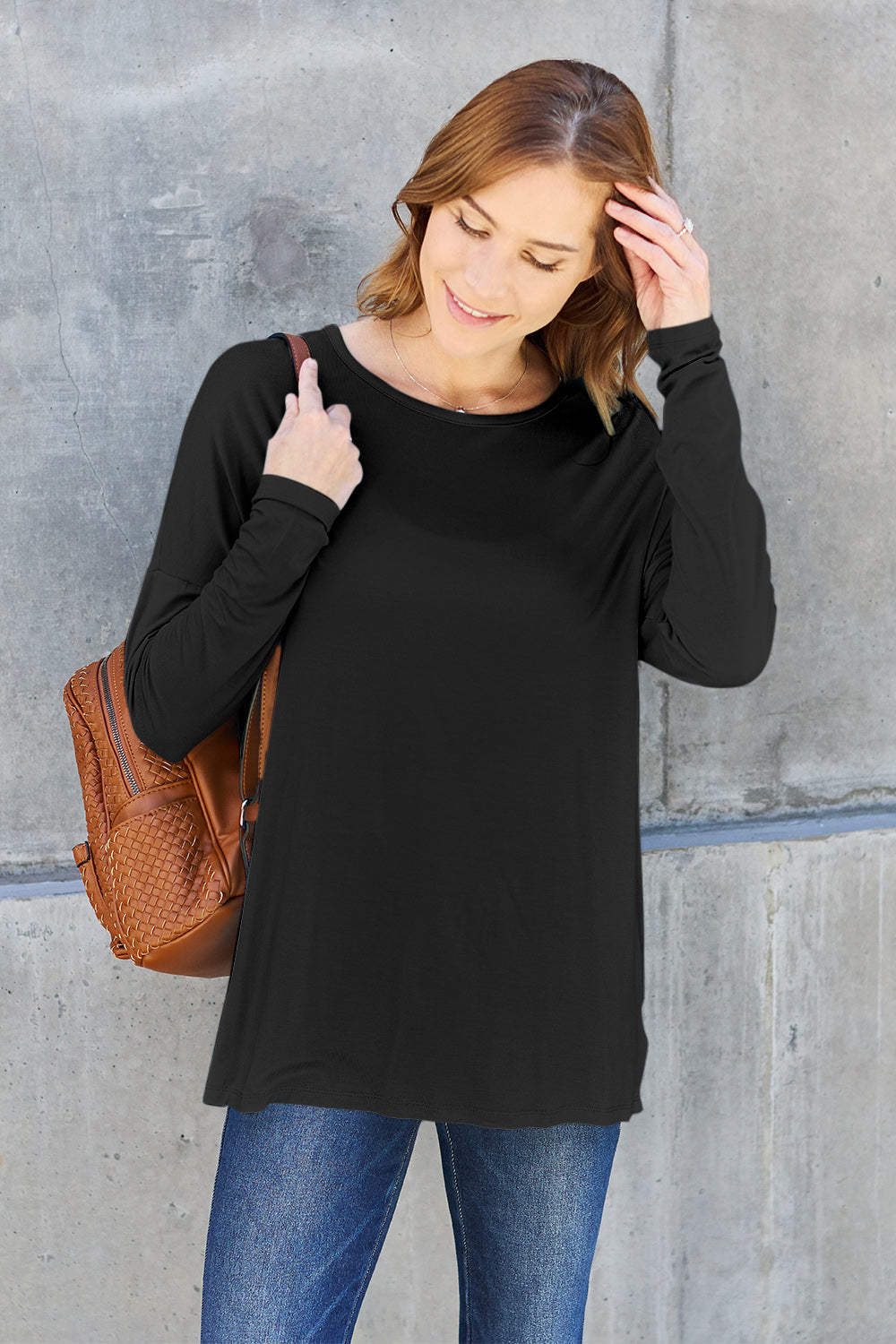 A woman with shoulder-length hair wearing a Basic Bae Full Size Round Neck Dropped Shoulder T-Shirt in green and a pair of jeans, exuding a basic style, stands against a concrete wall. She's holding an imported brown woven backpack on her shoulder while touching her hair with her hand.