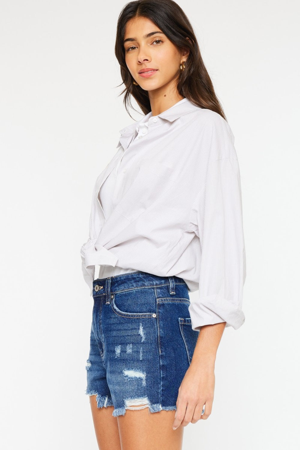 A woman with long dark hair wears a white blouse tied at the waist over a white top and Kancan High Rise Frayed Hem Denim Shorts, posing with one hand in her pocket against a plain background, embodying the perfect summer wardrobe.