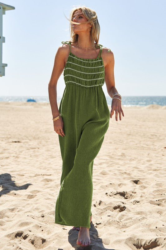 A person wearing the Washed Multi Smocked Detail Tie Straps Jumpsuit stands on the sunny beach, with the ocean creating a perfect backdrop.