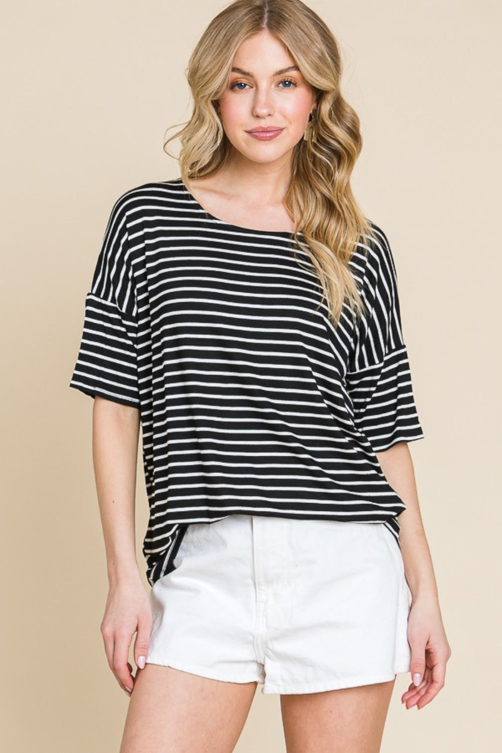 A woman with wavy blonde hair wears a BOMBOM Striped Round Neck T-Shirt and white shorts, standing against a beige background. This classic and casual look highlights her effortless style.